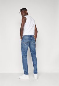 Τζιν παντελόνι σε μπλε απόχρωση, εφαρμοστό, με χαρακτηριστικές πίσω τσέπες και καφέ δερμάτινη λεπτομέρεια στη ζώνη, συνδυασμένο με μια αμάνικη λευκή μπλούζα και λευκά αθλητικά παπούτσια.