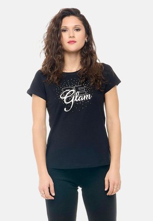 Jeune femme aux cheveux bouclés portant un t-shirt noir avec le texte « Glam » et des accents scintillants, debout devant un fond clair uni.