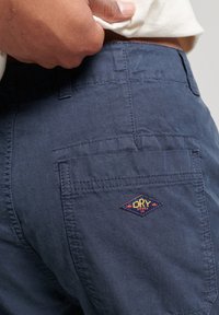 Pantalones cortos azul marino hechos de una tela texturizada, con un bolsillo trasero que presenta un detalle bordado en forma de diamante multicolor con el logo "DRY".