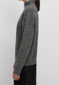 Donna di profilo che indossa un maglione grigio a collo alto e maniche lunghe, e una gonna nera, davanti a uno sfondo chiaro.