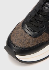 Detalhe do bico de um sapatênis castanho texturizado com acabamento em couro preto, cadarços pretos e sola acolchoada branca.