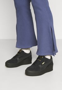 Černé semišové platformové tenisky s texturovanými detaily a zlatými akcenty, kombinované se širokými námořnickými kalhotami s bočními rozparky.