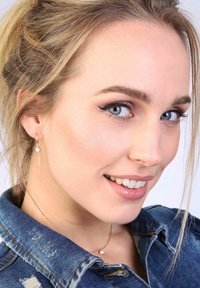 Gouden oorbellen met een hangend ontwerp en een kleine edelsteen. De persoon draagt een denim shirt met versleten details en een delicate gouden ketting.