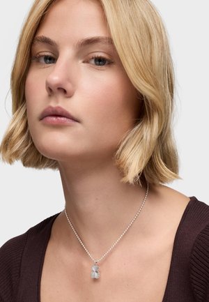 Collier en argent avec un design de feuille, fixé à une chaîne délicate. Le modèle porte un haut côtelé foncé sur un fond neutre.