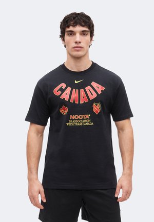 Mladý muž nosiaci čierne tričko s nápisom "CANADA" v červenej a žltej farbe, stojaci pred jednoduchým svetlým pozadím.