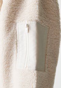 Lähikuva beige fleecetakista, jossa on sileä, tekstuuri kangas ja vetoketjullinen tasku kontrastivärisestä vaaleasta materiaalista hihassa.