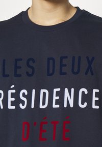 Námořnická modrá bavlněná trička s výrazným, barevným textem: "LES DEUX," "RÉSIDENCE," a "D'ÉTÉ" v bílé, červené a tmavě modré.