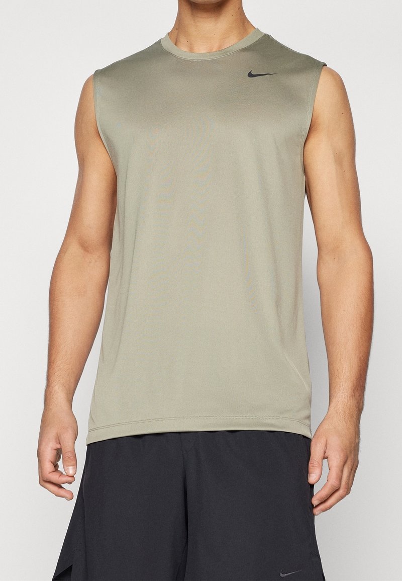 Homme portant un débardeur de sport vert olive clair et un short de sport noir, debout avec les bras détendus le long du corps devant un fond uni.