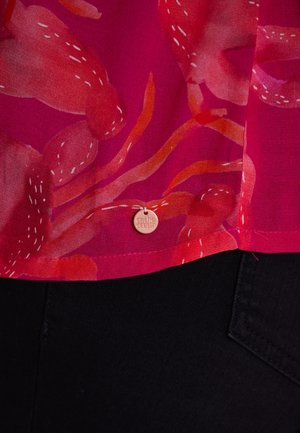 Blouse rose transparente avec des motifs floraux rouges et oranges ; comporte une petite étiquette en métal avec "cache cœur" près de l'ourlet. Tissu noir en dessous.