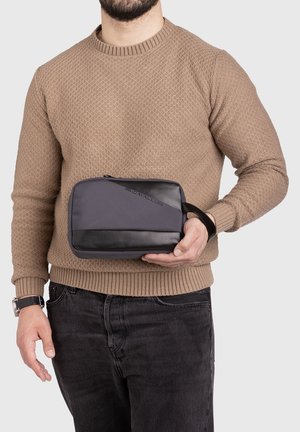 Homme en pull beige et jean noir tenant une pochette zippée grise et noire avec une sangle au poignet, sur fond blanc.