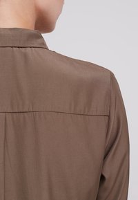 Camicia marrone con colletto, caratterizzata da una trama liscia, con design a yoke sul retro e vestibilità ampia, vista dalla spalla in giù.