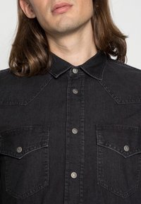 Czarna koszula dżinsowa z wyższym kołnierzem, srebrnymi zatrzaskami oraz dwiema kieszeniami na piersi z klapkami. Materiał ma gładką teksturę.
