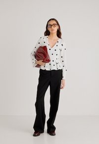 Hvit bluse med svarte polkadotter, V-hals, peplum-design; kombinert med svarte vide bukser og holder en rød clutch-veske.