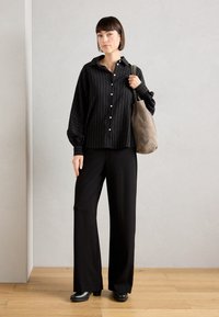 Chemise à rayures noires avec col, associée à un pantalon noir ample et des bottes noires épaisses. Sac fourre-tout en daim gris sur l'épaule.