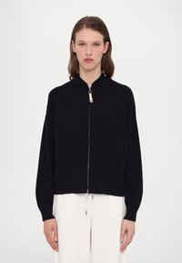 Sort strikket jakke med høy krage, løse ermer, frontlommer og detaljer i sølvglidelås. Glatt tekstur med et minimalistisk design.