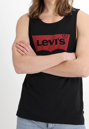 Osoba nosząca czarny bezrękawnik Levi's z czerwonym logo, z założonymi rękami i widocznymi na dole jasnoniebieskimi dżinsami.