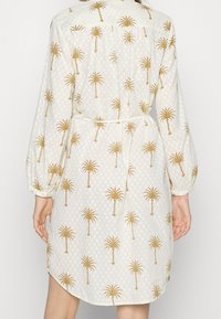 Vestido blanco con patrón de palmeras doradas, que presenta una tela texturizada, mangas largas y un cinturón atado en la cintura. Vista trasera visible.