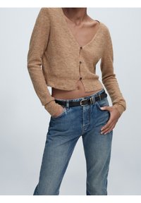 Hellbraune, kurz geschnittene Strickjacke mit V-Ausschnitt und drei Knöpfen, kombiniert mit blauen, gerade geschnittenen Jeans und einem schwarzen Gürtel mit silberner Schnalle.