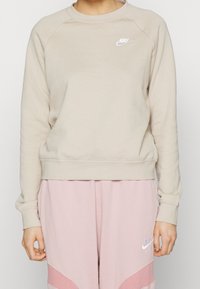 Beżowa bluza z polaru z rękawami raglanowymi, ściągaczami na mankietach i u dołu, z białym logo Nike na lewej piersi. Noszona z różowymi spodniami dresowymi.