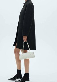 Sort uldfrakke med en struktureret silhuet, parret med en hvid lædertaske og sorte spidse sko. Teksturerede sorte sokker synlige.