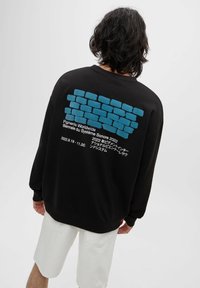 Czarna, oversize bluza z nadrukiem niebieskiego wzoru cegieł na plecach, z białym tekstem. Model ma na sobie białe szorty.