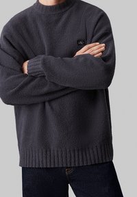 Ciemnoszary sweter z polaru z okrągłym dekoltem, ściągaczami przy mankietach i na dole, z małą czarną naszywką z logo na piersi.