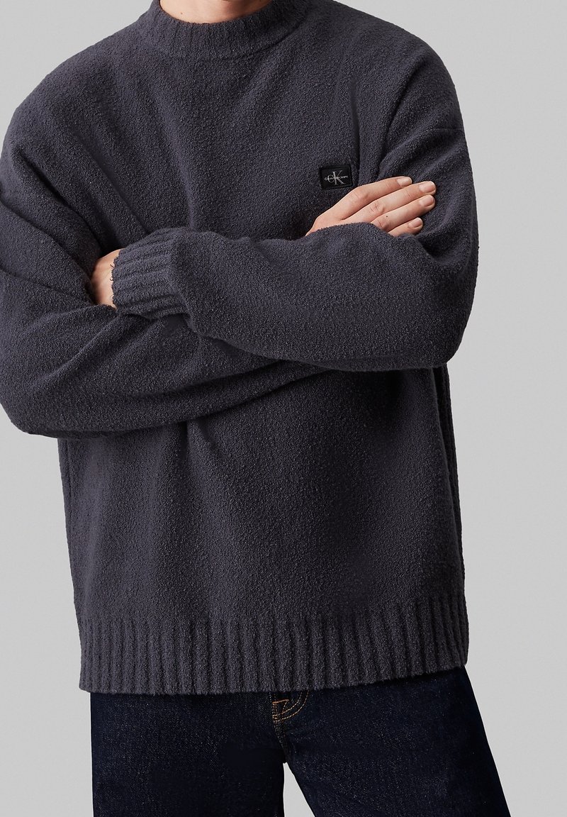 Ciemnoszary sweter z polaru z okrągłym dekoltem, ściągaczami przy mankietach i na dole, z małą czarną naszywką z logo na piersi.