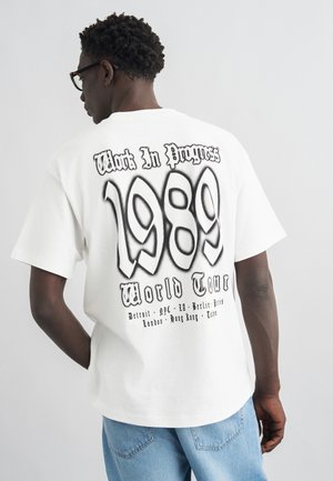 Bărbat purtând un tricou alb cu text negru „Work In Progress 1989 World Tour” și numele orașelor pe spate, asortat cu blugi albastru deschis.