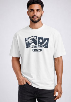 Man wearing white T-shirt with black graphic of stylized ocean waves, sun, and Mount Fuji, and text "TOKYO" on front.