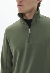 Pullover verde oliva con zip a un quarto in tessuto morbido, dotato di colletto alto e chiusura con zip, cuciture semplici e una vestibilità comoda.