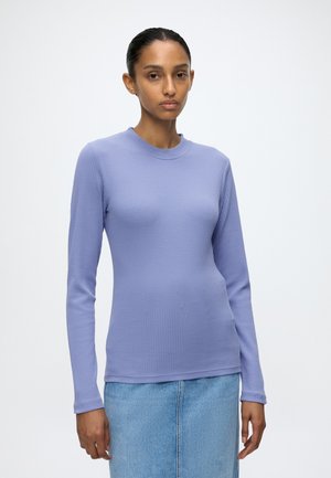 Femme aux cheveux foncés attachés en arrière portant un haut ajusté à manches longues violet clair et une jupe en jean bleu clair sur un fond uni.