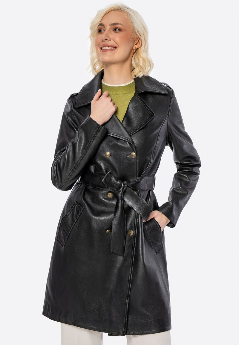 Donna con i capelli biondi che indossa un trench in pelle nera con cintura sopra una maglietta verde, sorridente e posando con una mano sul colletto.