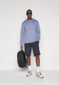 Μακρύ μανίκι ελαφρού μωβ πουκάμισου με υφή, συνδυασμένο με σκούρα σορτσάκια, λευκές κάλτσες και γκρι αθλητικά παπούτσια. Μαύρο σακίδιο κρατείται στο χέρι.