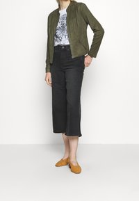 Chaqueta bomber de ante verde oliva con cremallera frontal, combinada con jeans negros ajustados y mocasines de mostaza tejidos. Camiseta gráfica debajo.