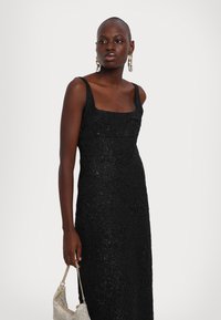 Schwarzes ärmelloses Kleid mit quadratischem Ausschnitt, strukturiertem Stoff, figurbetonter Silhouette und dezentem Schimmer, kombiniert mit einer metallischen Handtasche.