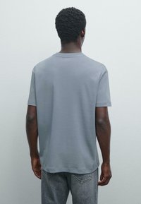 Šedé bavlněné tričko s kulatým výstřihem a krátkým rukávem, s volným střihem. Záda ukazují minimální švy a hladkou texturu.