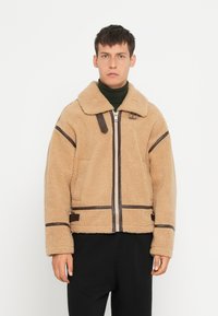 Beige shearling jakke med brun læderdetalje, lynlås lukning og en bred krave. Designet har en boxy form og er fuldt forret. 
