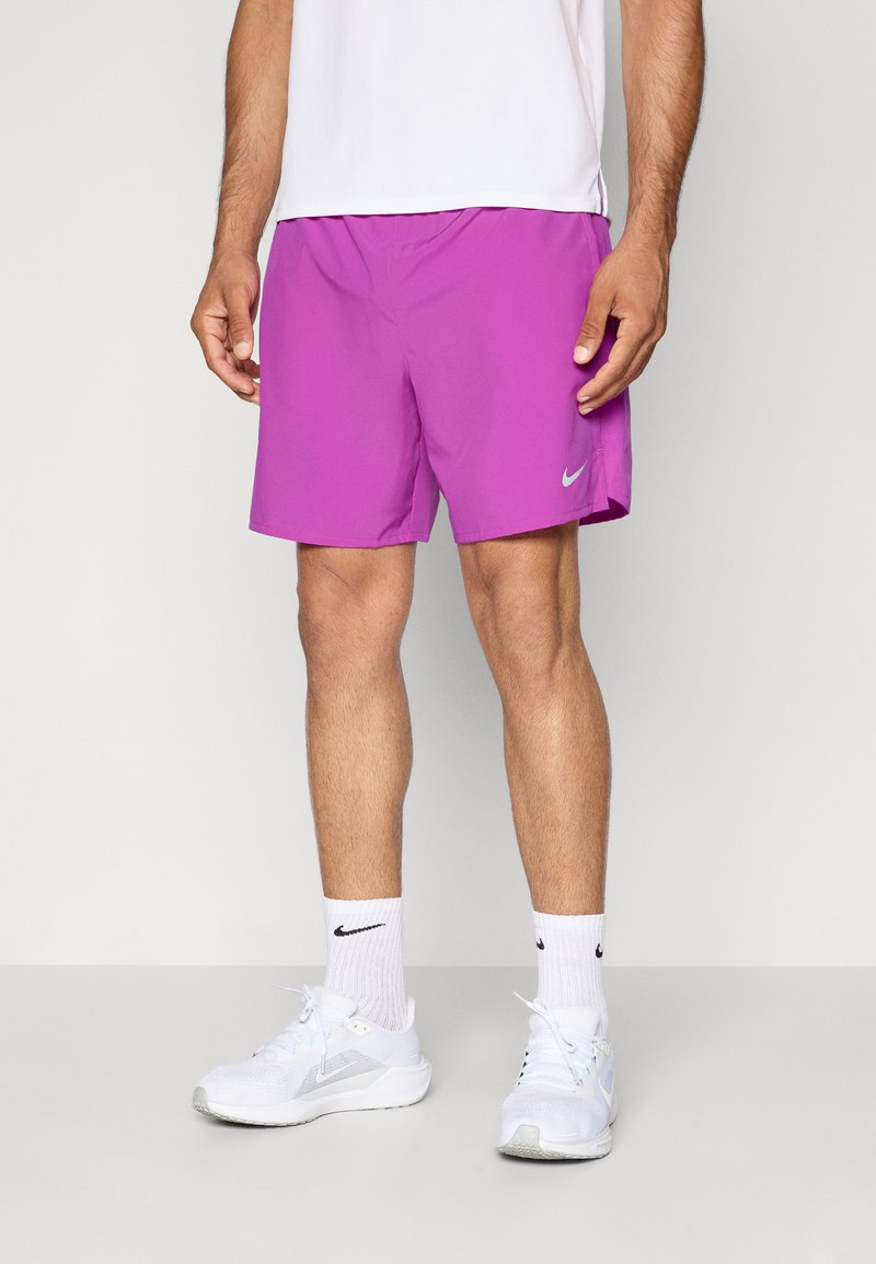 Homme portant un short de sport violet vif, des chaussettes blanches Nike et des chaussures de course blanches, se tenant devant un fond clair.