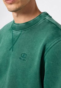 Hombre vistiendo un sudadera de cuello redondo verde con el logo de Champion bordado en el lado izquierdo del pecho, contra un fondo liso.