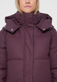 Donna che indossa una giacca imbottita viola con collo alto e dettagli con bottoni a pressione sul cappuccio, rivolta in avanti su uno sfondo semplice.