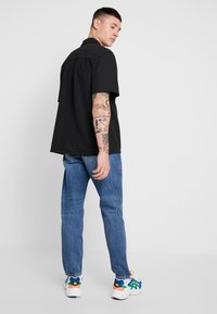 Camisa preta de colarinho com mangas curtas, combinada com jeans azul claro e sapatilhas coloridas. Tatuagens visíveis nos braços.