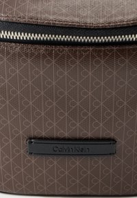 Καφέ ανάγλυφο υλικό με γεωμετρικό σχέδιο, με μαύρο φερμουάρ και ορθογώνια μαύρη ετικέτα με το κείμενο "Calvin Klein".