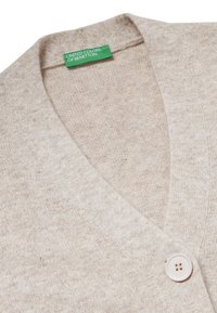 Cárdigan beige hecho de tela de punto texturizada, con escote en V y un solo botón de color claro. La etiqueta dice "United Colors of Benetton."