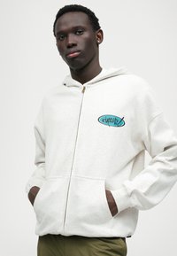 Helehallide tõmbluku hoodie pehmest kangast, millel on kappa, küljetaskud ja türkiissinine graafiline logo vasakul rinnal.