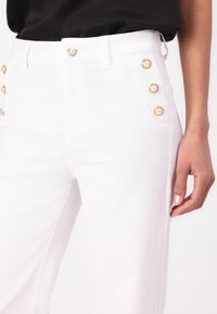 Pantaloni in denim bianco con vita alta, caratterizzati da bottoni dorati decorativi sulle tasche laterali e una texture morbida.