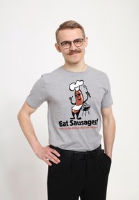 Camiseta de algodón gris con un chef de salchichas de dibujos animados sosteniendo una espátula, con un texto en negrita que dice "¡Come Salchichas!" en negro y naranja.