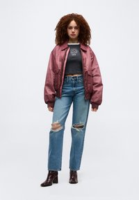 Γυναίκα με κατσαρά μαλλιά που φοράει ροζ μπουφάν τύπου bomber, μαύρο κοντό τοπ με γραφικά, σκισμένο μπλε τζιν και σκούρα μπότες με τακούνια, στέκεται μπροστά σε λευκό φόντο.