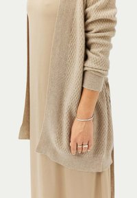 Beige stickad cardigan med strukturerad mönster, öppen front och ribbade manschetter, bärs över en beige klänning. Enkel silverarmband på handleden.