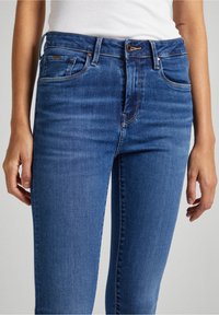 Jeans de mezclilla azul con un diseño de cintura alta, estilo de cinco bolsillos y sutiles desgastes a lo largo de las piernas. Textura suave y corte ajustado.