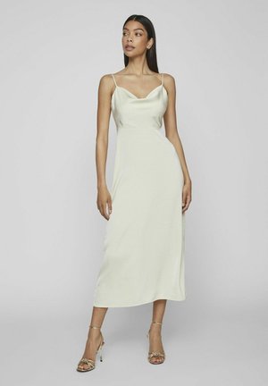 Femme portant une robe midi sans manches couleur crème avec de fines bretelles et des sandales dorées à talons hauts, debout devant un fond clair et uni.